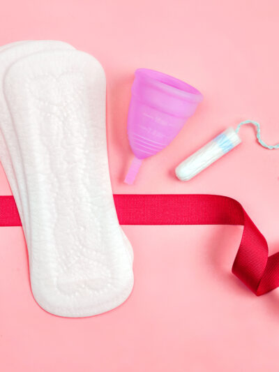 Sanitary pads, a menstrual cup, and a tampon sits on a pink background. A red ribbon flows around these products.