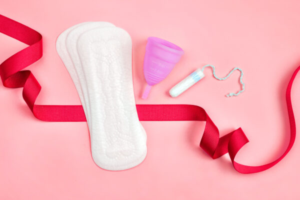 Sanitary pads, a menstrual cup, and a tampon sits on a pink background. A red ribbon flows around these products.