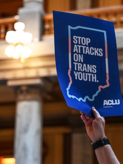 Inside the Indiana statehouse, a hand holds up a protest poster that reads, "stop attcks on trans youth"