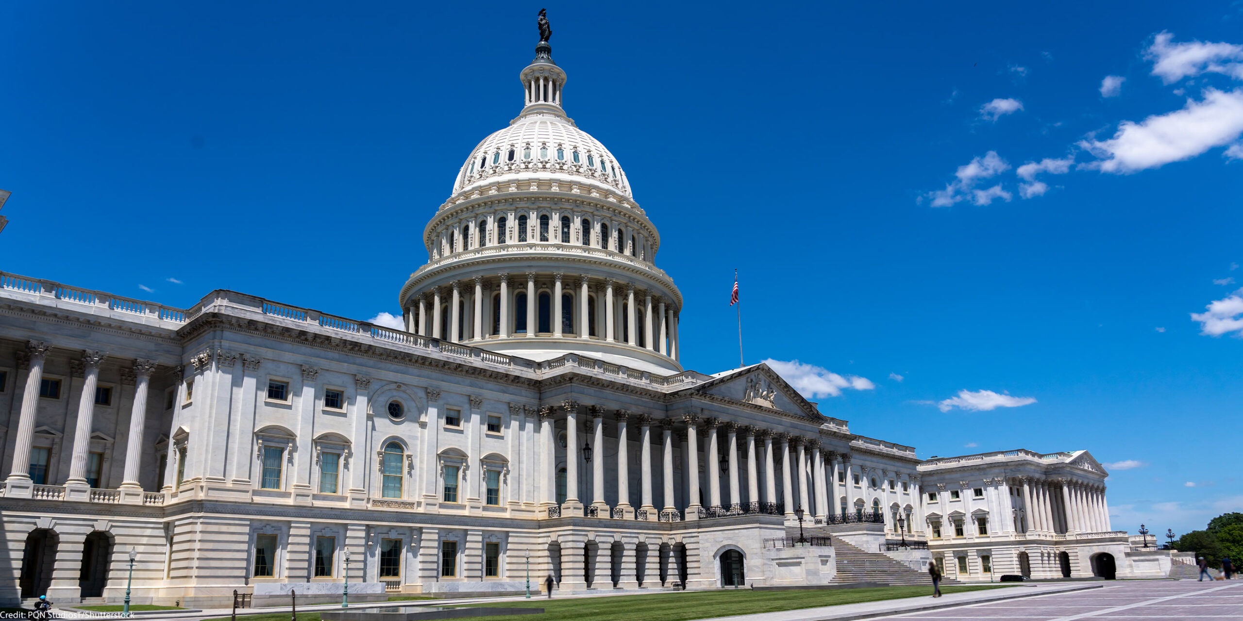 Wide daytime view of the U.S. Capitol building in Washington, D.C., with the white dome and columns under a bright blue sky.