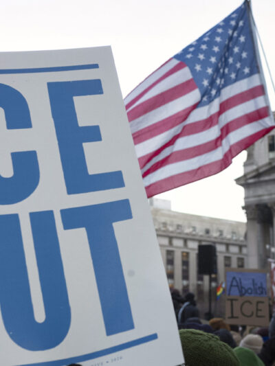 A hand holds a large white protest sign reading "ICE OUT" in bold blue letters during a crowded demonstration outside a government building. An American flag waves above the crowd, and additional signs in the background include messages calling to abolish ICE.