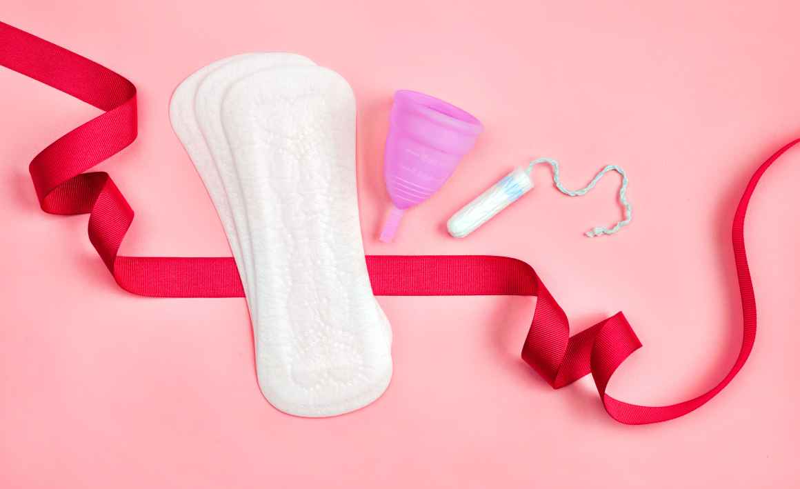 Sanitary pads, a menstrual cup, and a tampon sits on a pink background. A red ribbon flows around these products. 