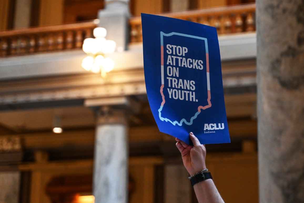 Inside the Indiana statehouse, a hand holds up a protest poster that reads, "stop attcks on trans youth" 