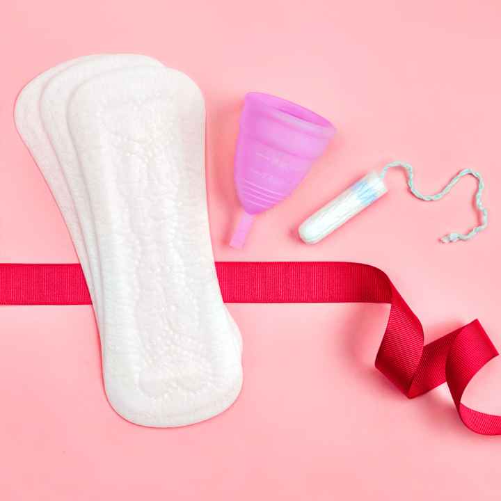 Sanitary pads, a menstrual cup, and a tampon sits on a pink background. A red ribbon flows around these products.