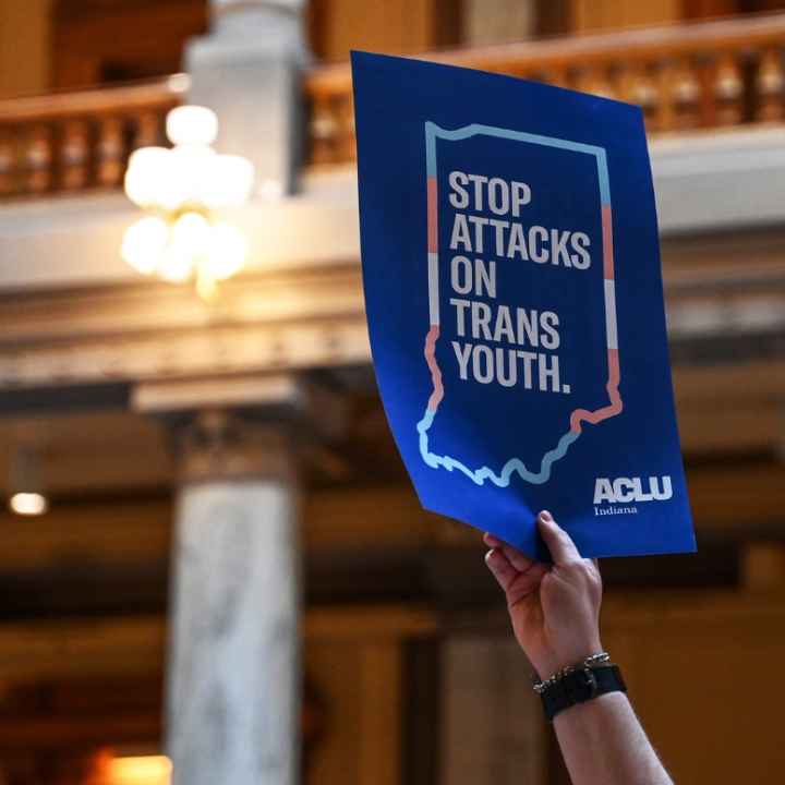 Inside the Indiana statehouse, a hand holds up a protest poster that reads, "stop attcks on trans youth"